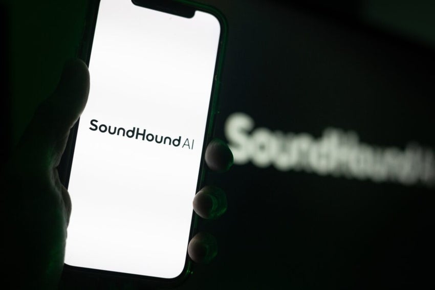 Silhouette of Hand Holding iPhone with SoundHound AI Logo in Front of Computer Screen Displaying Logo