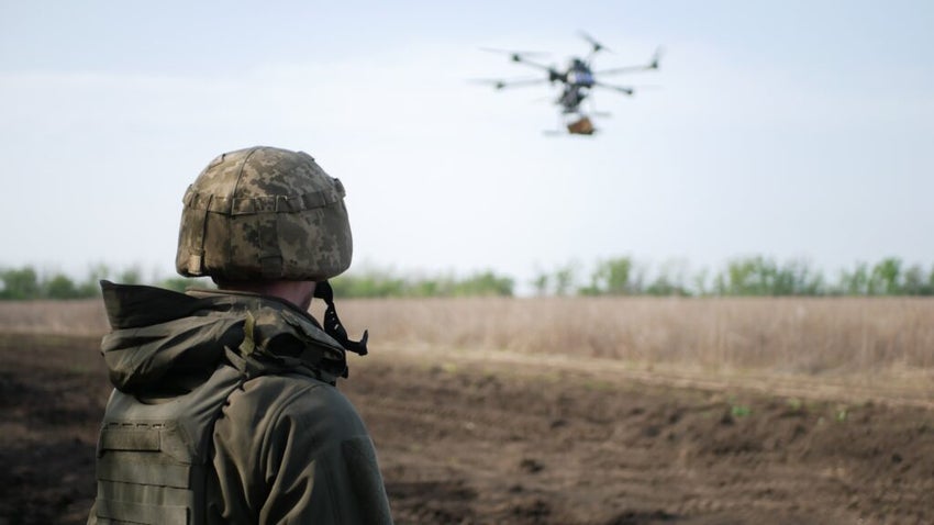 A Soldier In Camouflage Observes A Drone Hovering Above An Open Field