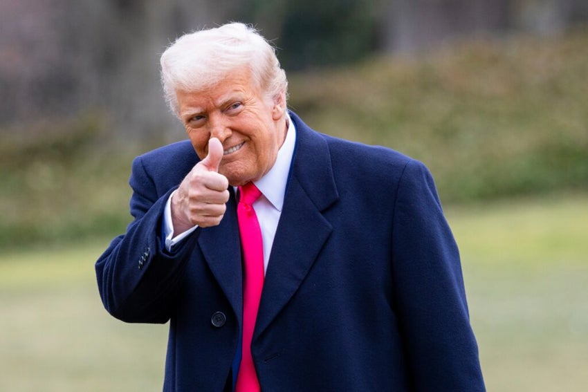 President Donald Trump in a dark coat and bright pink tie gives a thumbs-up gesture outdoors with a blurred green background.